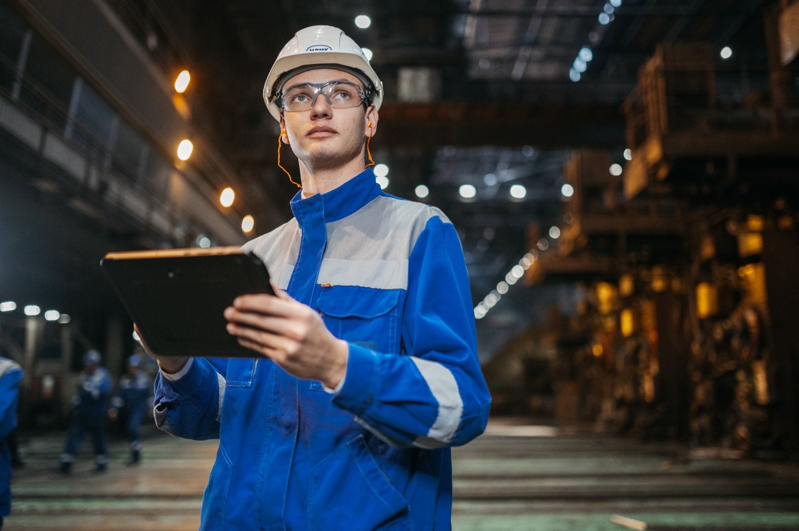 A plant worker in a hard hat and safety glasses holding a tablet on a steel production floor