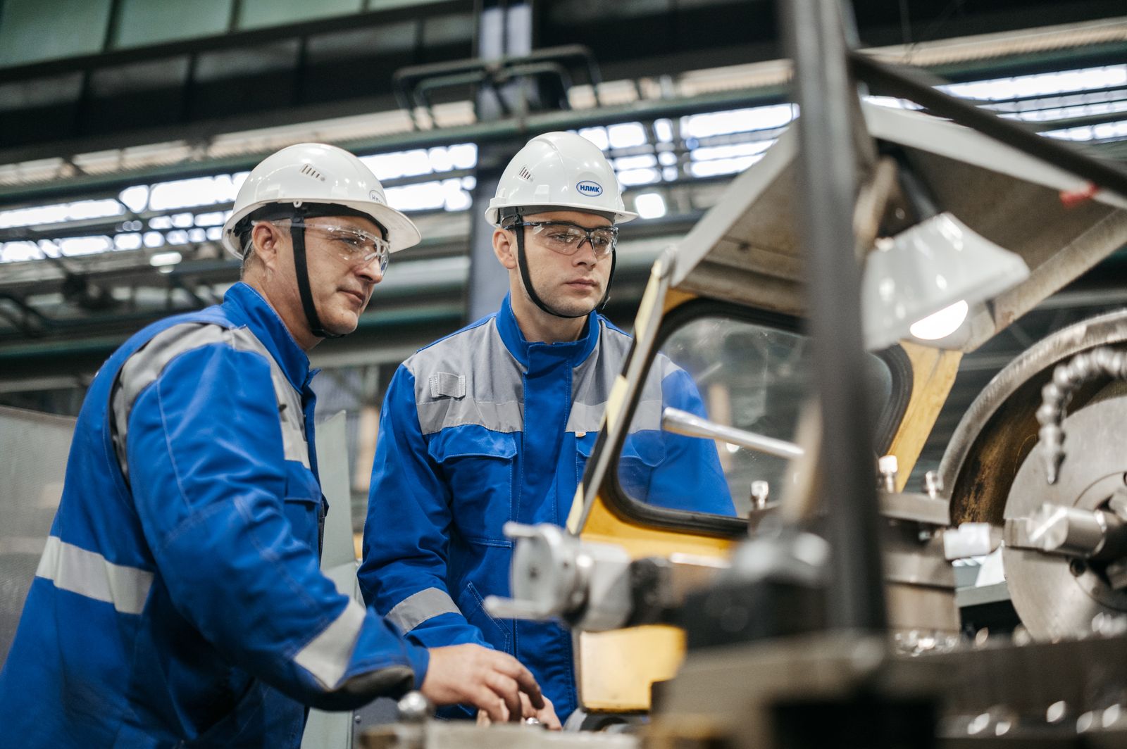An experienced maintenance technician and a younger one in full PPE working together at a machine